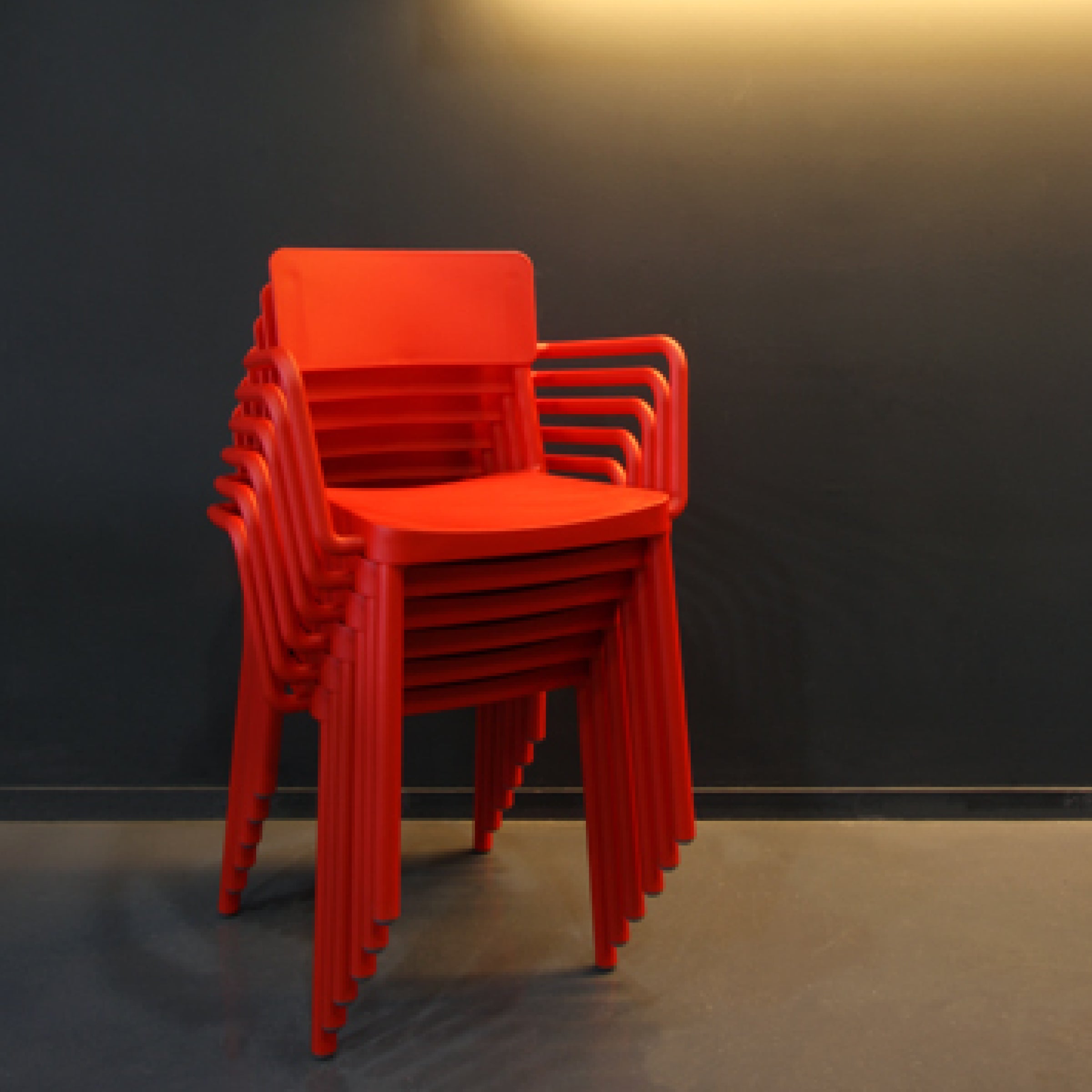 A stack of Lisboa Armchairs from Resol is neatly arranged against a dark wall. The top chair is fully visible, bathed in a warm light that contrasts with the background. Crafted sustainably, the floor beneath is smooth and even.