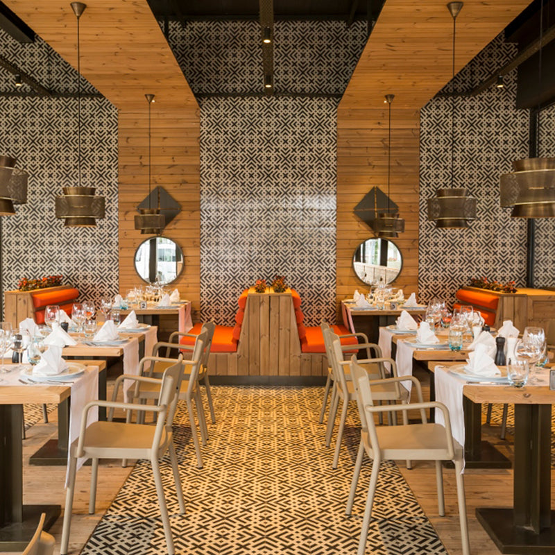 The stylish restaurant boasts patterned black and white walls and flooring, equipped with Resols Lisboa Armchairs. Wooden tables are set with white napkins and glasses, while round mirrors, orange-cushioned seating, and metal hanging lights enhance the ambiance.