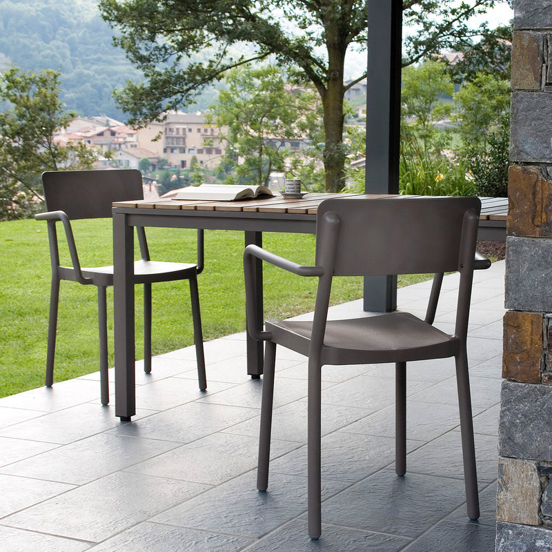 The patio showcases two brown Lisboa Armchairs and a wooden table from Resol Designer Furniture on a tiled floor, with a book and mug on the table; the scene is set against a backdrop of green lawn, trees, and UV-protected hills dotted with houses.