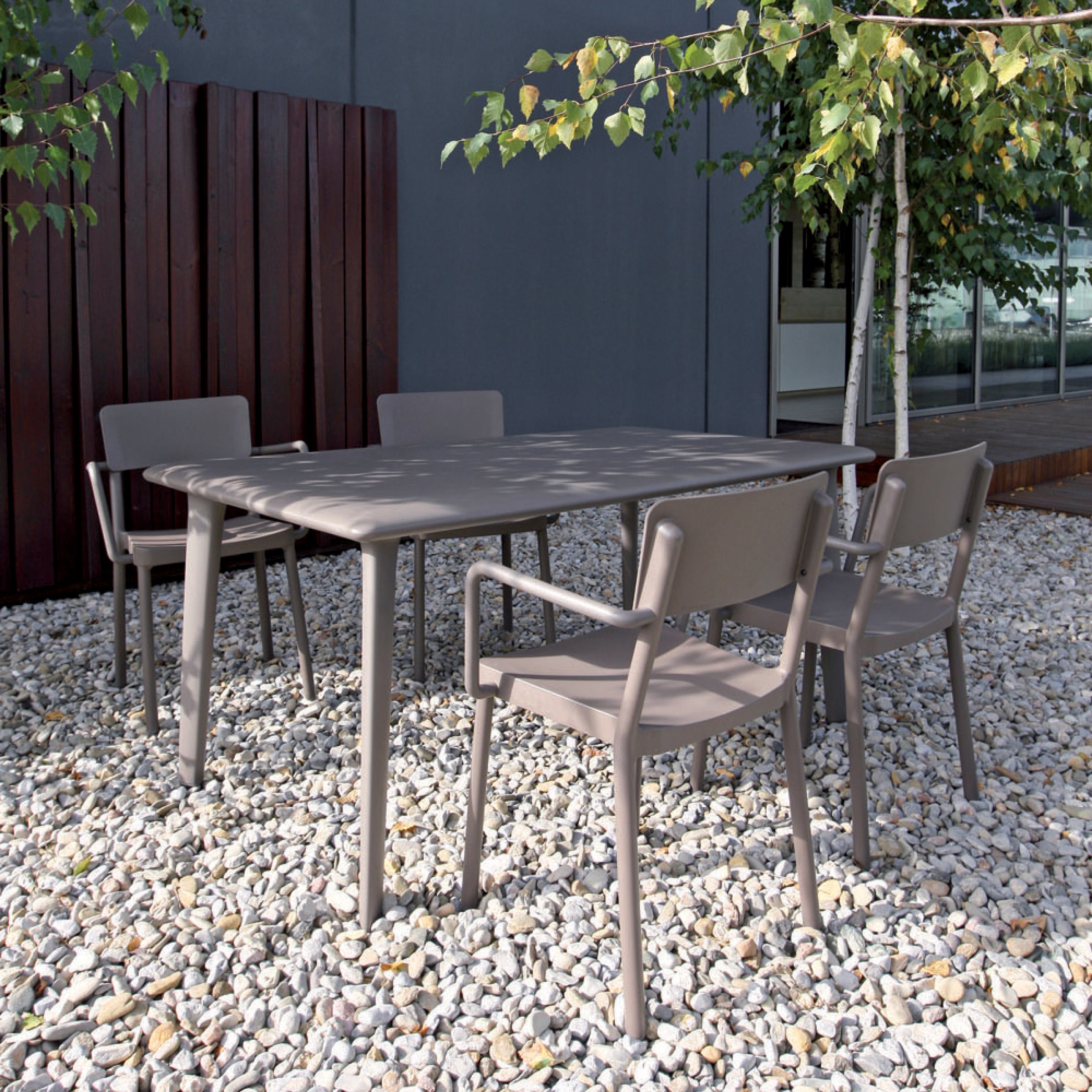 Outdoor patio setting featuring Resols Lisboa Armchair, a rectangular table, and four matching chairs on a pebble-strewn surface. Background includes a wooden fence, leafy trees, and a modern building with large windows—all crafted sustainably.