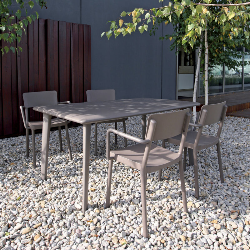 Outdoor patio setting featuring Resols Lisboa Armchair, a rectangular table, and four matching chairs on a pebble-strewn surface. Background includes a wooden fence, leafy trees, and a modern building with large windows—all crafted sustainably.