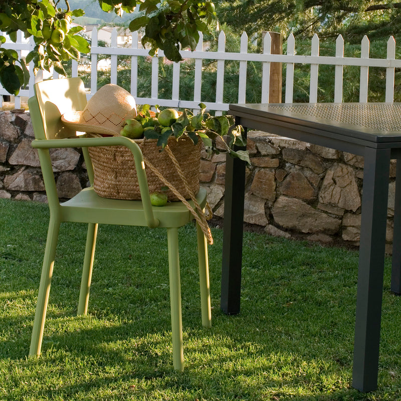 The Lisboa Armchair in green from Resol stands on grass, showcasing sustainable design. It supports a woven basket of apples and a straw hat beside a table. Behind it, trees rise against a stone wall and white picket fence.