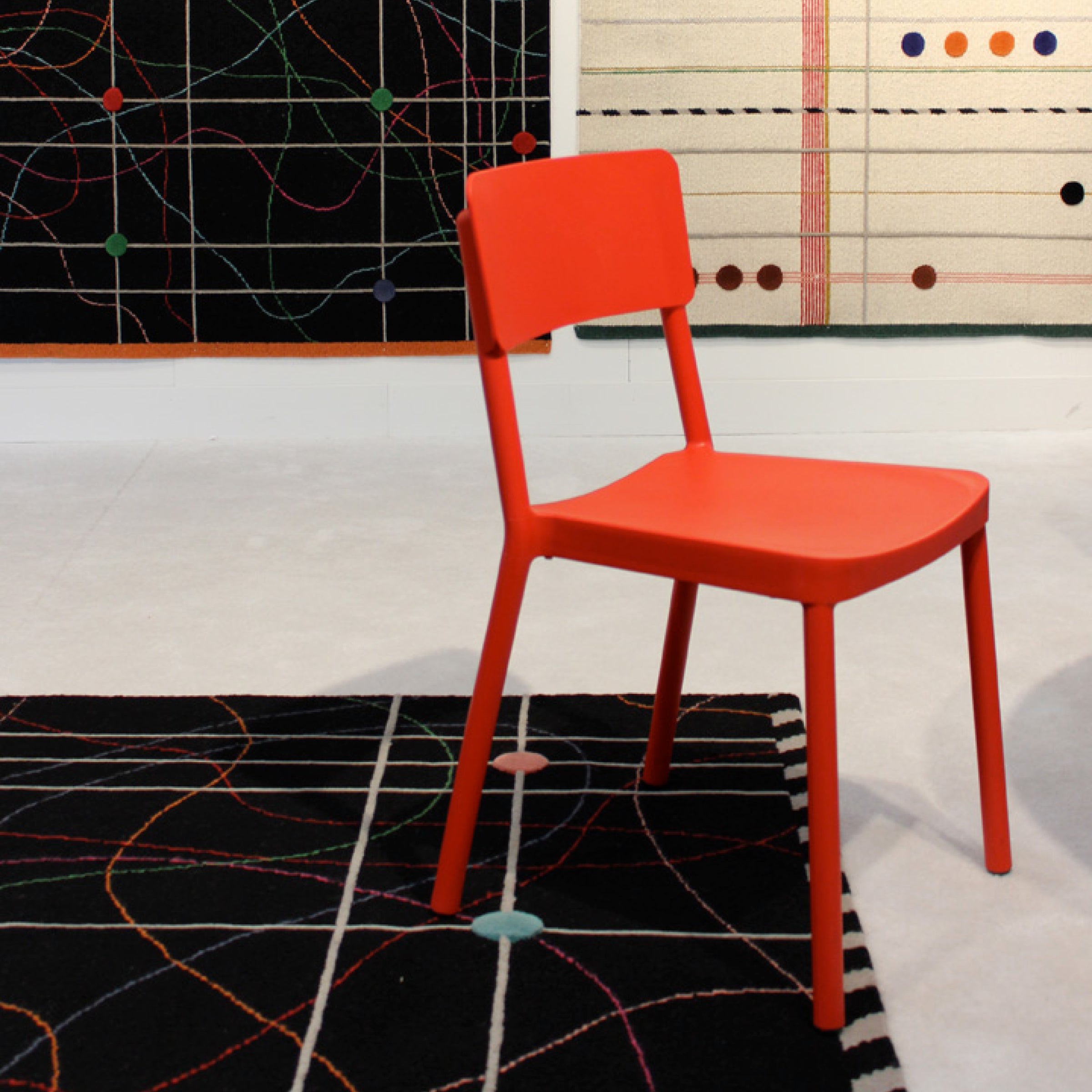 A Lisboa Chair by Resol, in red, sits on a patterned rug in front of a wall decorated with abstract geometric art. The rug and UV-protected artwork feature complementary designs of intersecting lines and colorful dots, capturing modern minimalism.