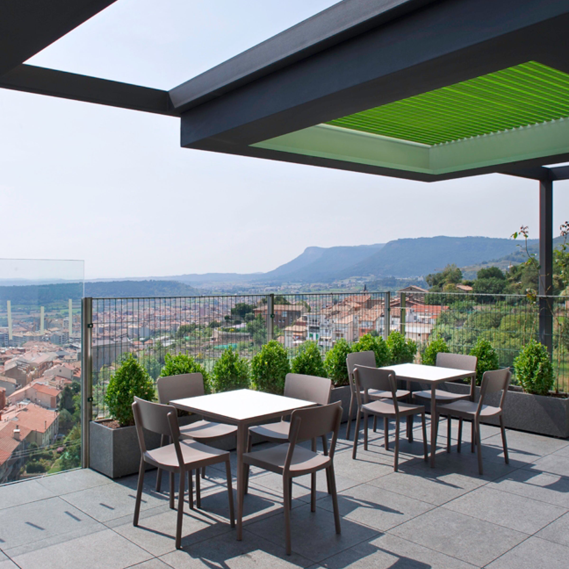 The outdoor terrace features Resols Lisboa chairs, with tables perfectly arranged to overlook a scenic view of the town and mountains. The area is shaded by a modern pergola offering UV protection, while green plants and glass railings add an elegant touch.