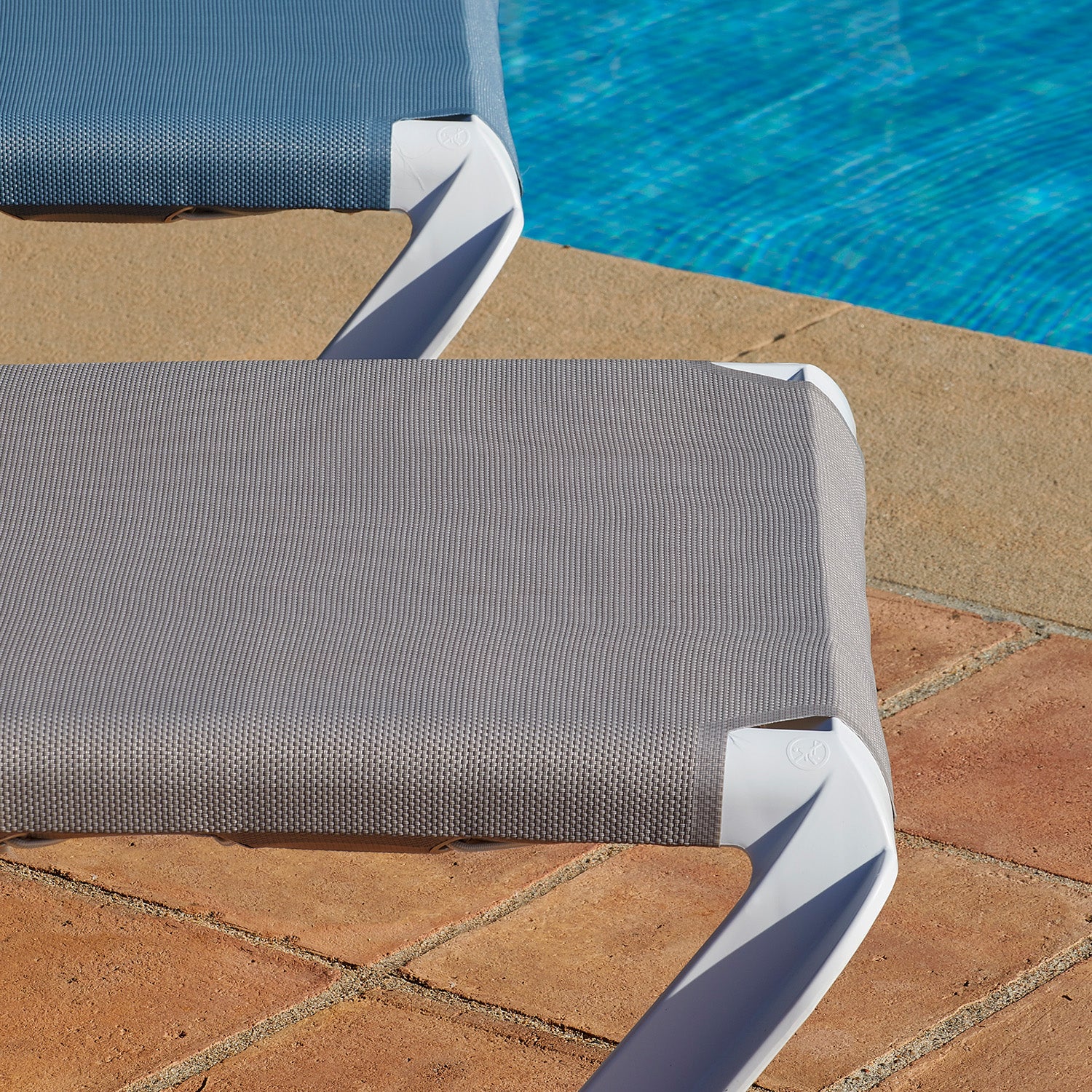 Two gray Marina Club Sunloungers by Resol with white frames are reclined by a bright blue swimming pool on a terracotta-tiled patio. Sunlight casts subtle shadows across the scene, creating an inviting lounging spot.