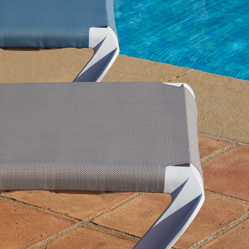 Two gray Marina Club Sunloungers by Resol with white frames are reclined by a bright blue swimming pool on a terracotta-tiled patio. Sunlight casts subtle shadows across the scene, creating an inviting lounging spot.
