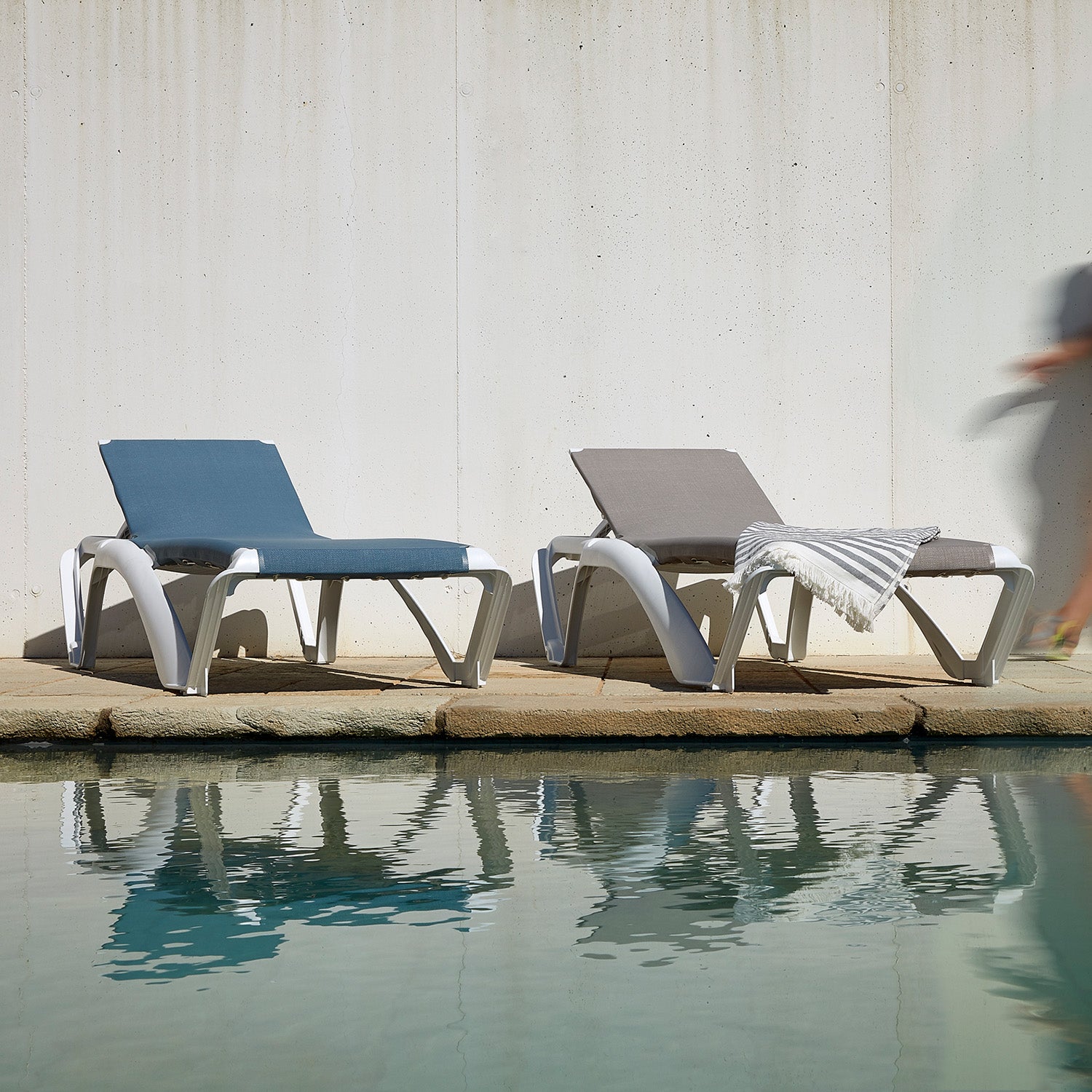 Two Marina Club Sunloungers from Resol, one blue and one gray with a striped towel, are reclinable and set by a tranquil pool. A white concrete wall forms the backdrop, while a blurred figure walks past on the right.