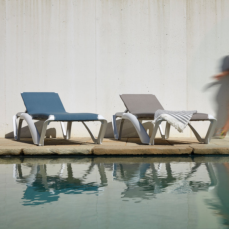 Two Marina Club Sunloungers from Resol, one blue and one gray with a striped towel, are reclinable and set by a tranquil pool. A white concrete wall forms the backdrop, while a blurred figure walks past on the right.