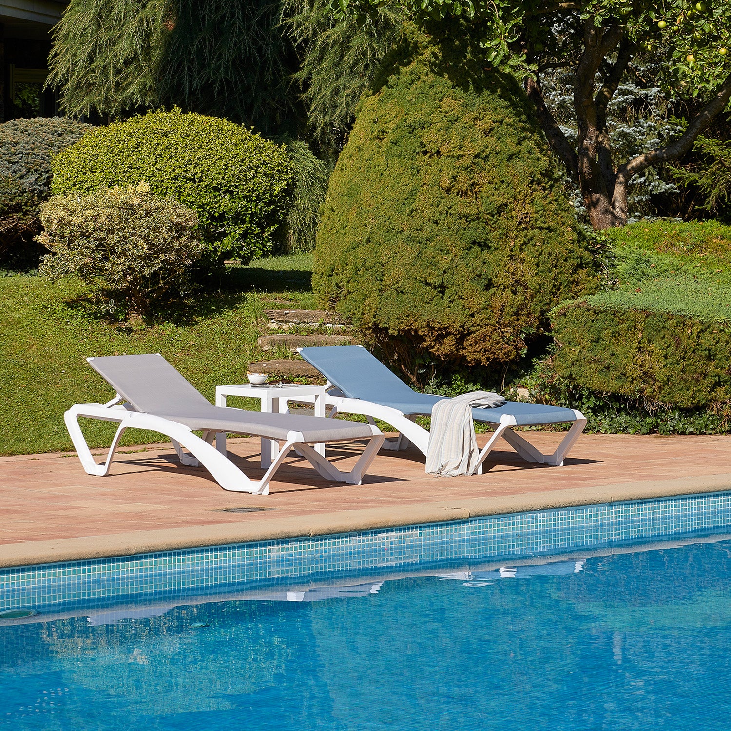 Two white Marina Club Sunloungers by Resol, featuring gray cushions and reclinable positions, are placed next to a blue pool on a brick patio. A small white table enhances the setup, with a sustainably maintained garden flourishing under the clear sky in the background.