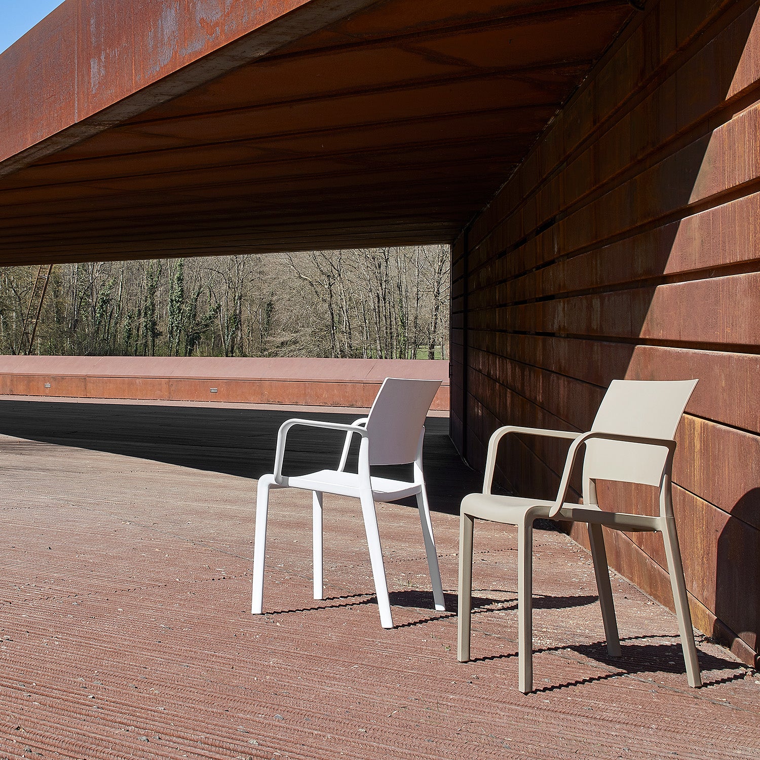 Two modern Fiona Armchairs by Resol, one white and one beige, are stacked on a textured surface next to a rust-colored metal wall beneath an overhang. Bare trees are visible in the background.