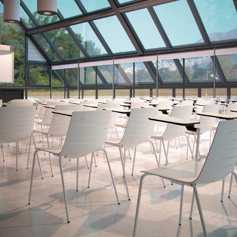 A modern, sunlit classroom features Resols white Skin Chairs with attached desks, made from eco-friendly fiberglass and polypropylene. The polished floor enhances the glass walls with green views, while slanted windows fill the room with natural light.