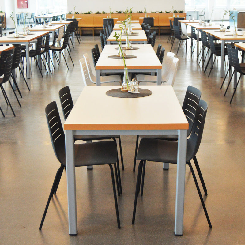 The spacious cafeteria is furnished with rows of tables and Resols black Skin Chairs, crafted from sustainable fiberglass and polypropylene. Each table has a centerpiece with a vase and plant. Bright lighting and a neutral color scheme create an airy, inviting atmosphere.