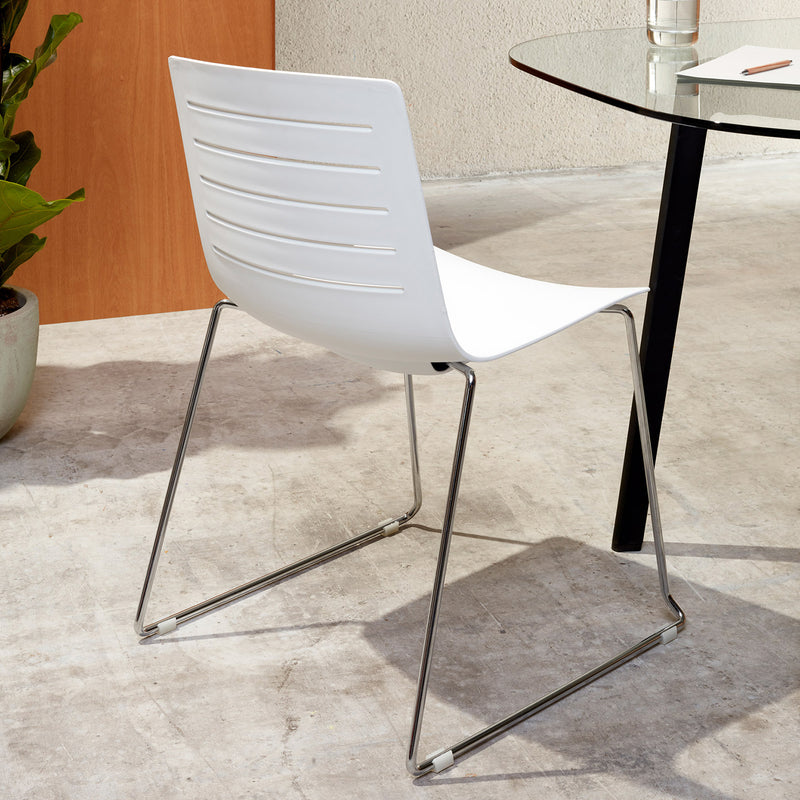 The Resol Skin Sled Chair, featuring a modern design with fiberglass and polypropylene seat and metal legs in white, is placed beside a transparent glass table on a concrete floor. A small potted plant and wooden panel complement the sustainable furniture theme in the background.