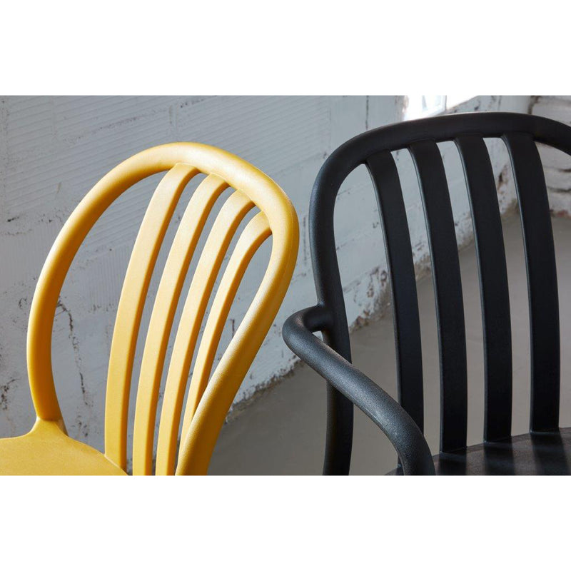 Close-up of two innovative Resol Sue Chairs, one yellow and one black, featuring vertical slatted backs. Positioned side by side on a concrete floor against a textured white wall, they exemplify the blend of style and sustainable production.