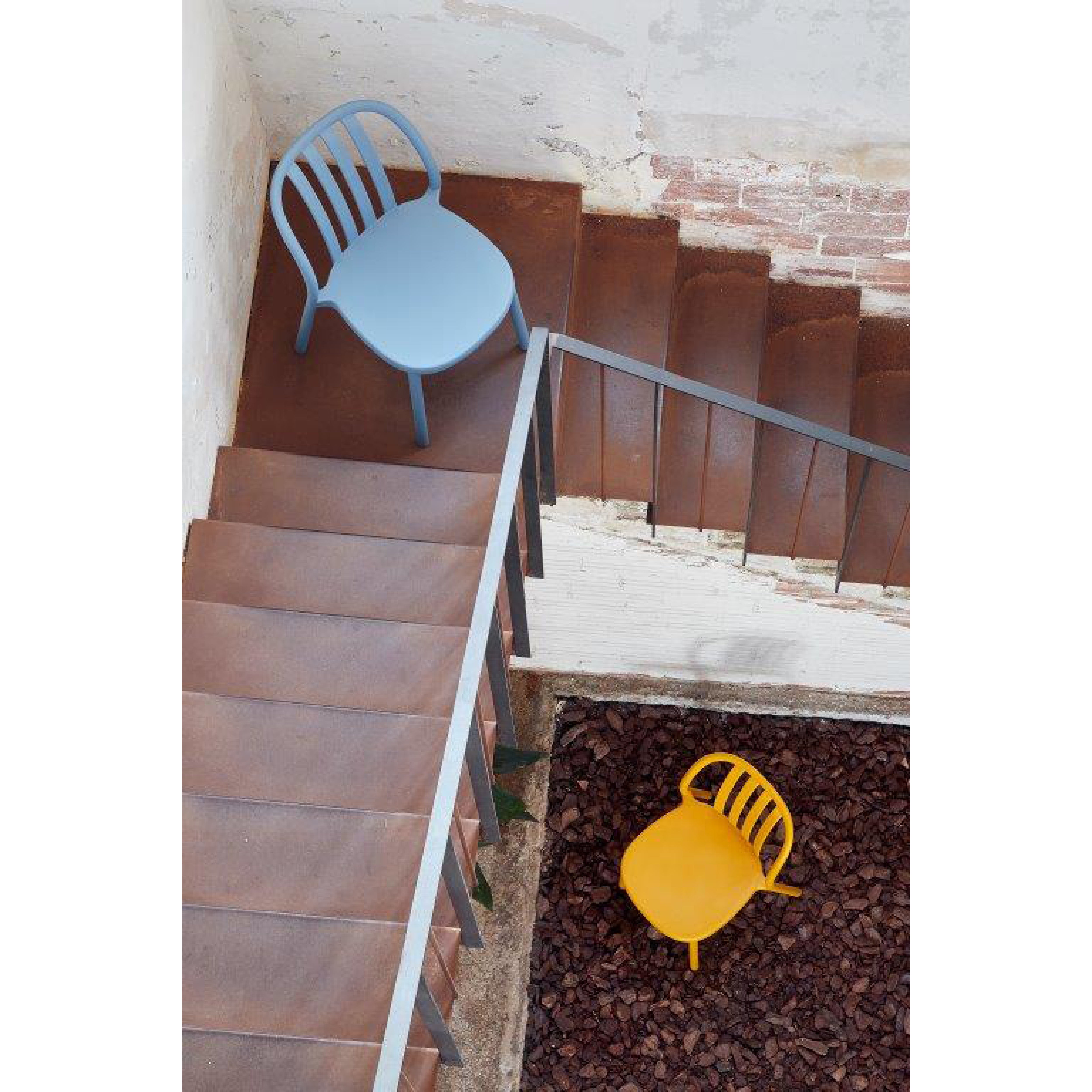 A staircase leads to a landing with a light blue Resol Sue Chair, featuring a rusted metal floor and railing. Below, a yellow chair rests on the rocky ground. The surroundings offer a rustic, industrial style suggestive of sustainable production methods.