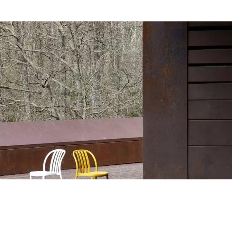 Two empty Sue Armchairs by Resol, one white and one yellow, crafted sustainably, sit on an outdoor patio near a rust-colored railing. In the background, leafless trees contrast with a large dark structure on the right.