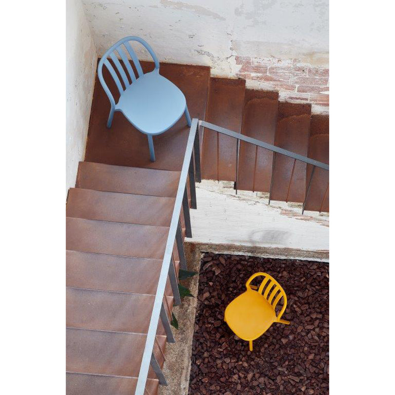 A staircase leads to a landing with a light blue Resol Sue Chair, featuring a rusted metal floor and railing. Below, a yellow chair rests on the rocky ground. The surroundings offer a rustic, industrial style suggestive of sustainable production methods.