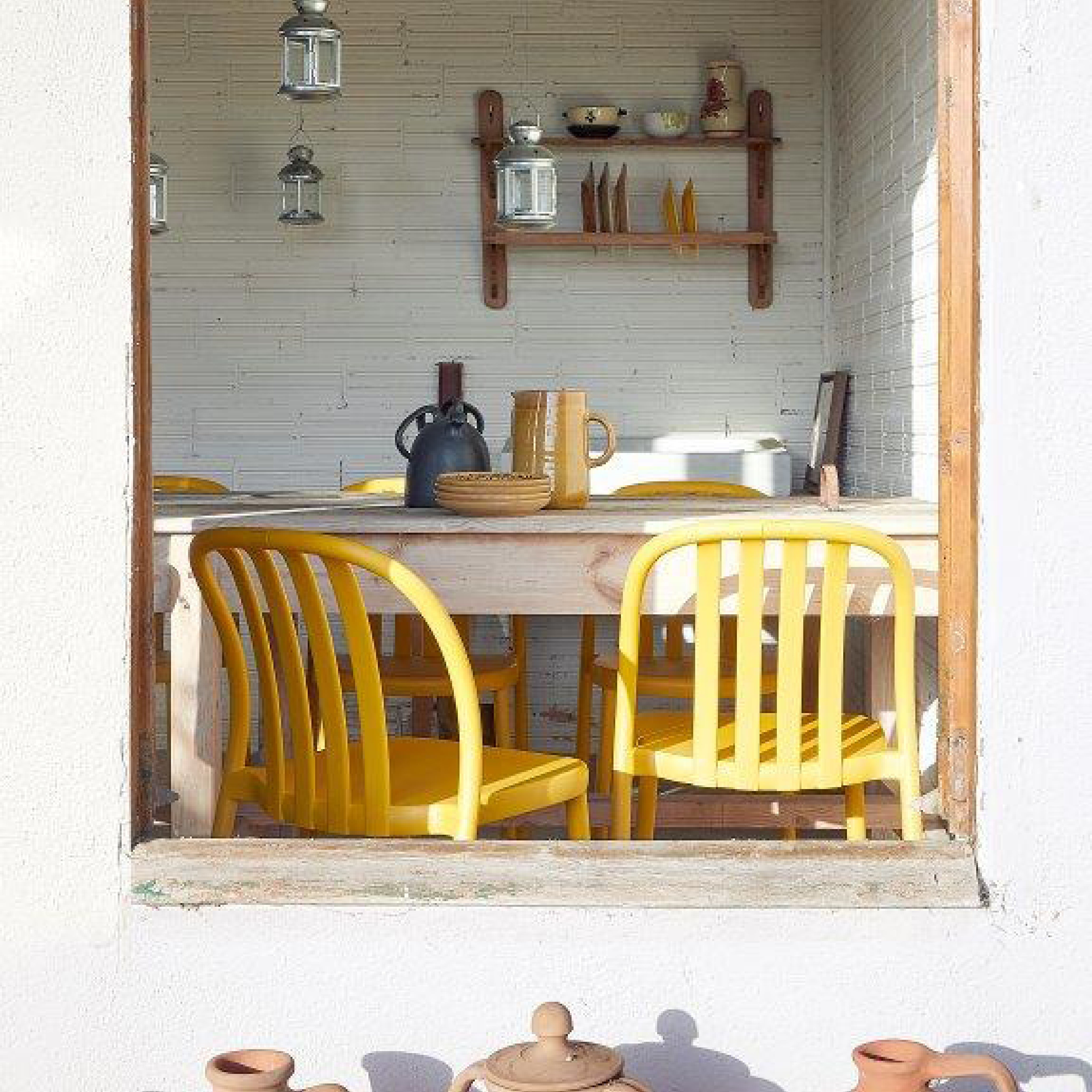 Through a window, a cozy dining area reveals a wooden table with a brown pitcher and basket. Surrounding it are four Sue Chairs by Resol, crafted from innovative furniture practices. A wooden shelf with jars and bowls adorns the back wall, and terracotta pots rest on the windowsill.