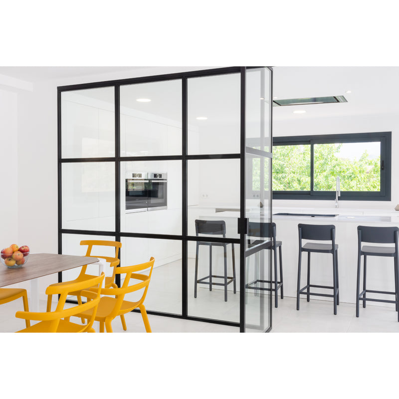 The modern kitchen showcases white cabinetry and a chic black metal-framed glass partition. A sustainably produced built-in oven gleams nearby. On the left, a wooden dining table is paired with Resols Tokyo yellow chairs, while three black bar stools sit by the white island near a large window.