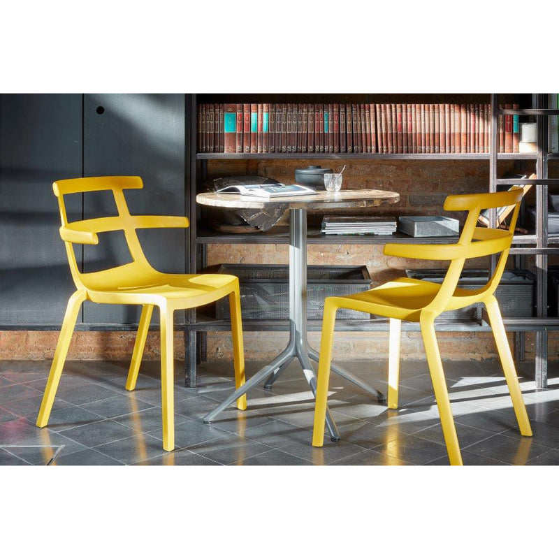 Two modern Tokyo chairs from Resol flank a small, round marble-topped table holding a book and glass. This setup sits against a brick wall adorned with bookshelves, all on a stylish tiled floor.