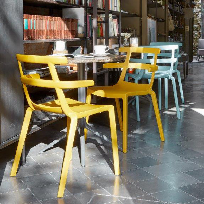 In a cozy cafe, a round table is surrounded by Resols Tokyo Chairs in vibrant colors—two yellow and one blue. UV-protected sunlight creates patterns on the tiled floor as books fill the shelves, and a cup with saucer sits invitingly on the table.