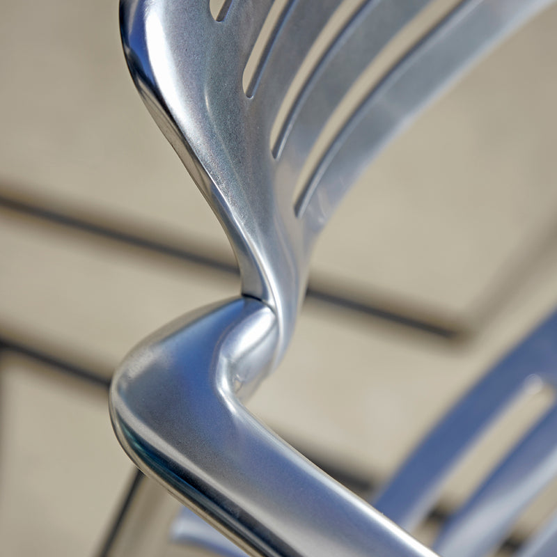 The Toledo Armchair by Resol shines with a sleek metallic finish, featuring a twisted design and evenly spaced slats on the back, suggesting its sustainable production. The background is softly blurred, highlighting the chairs elegant details.