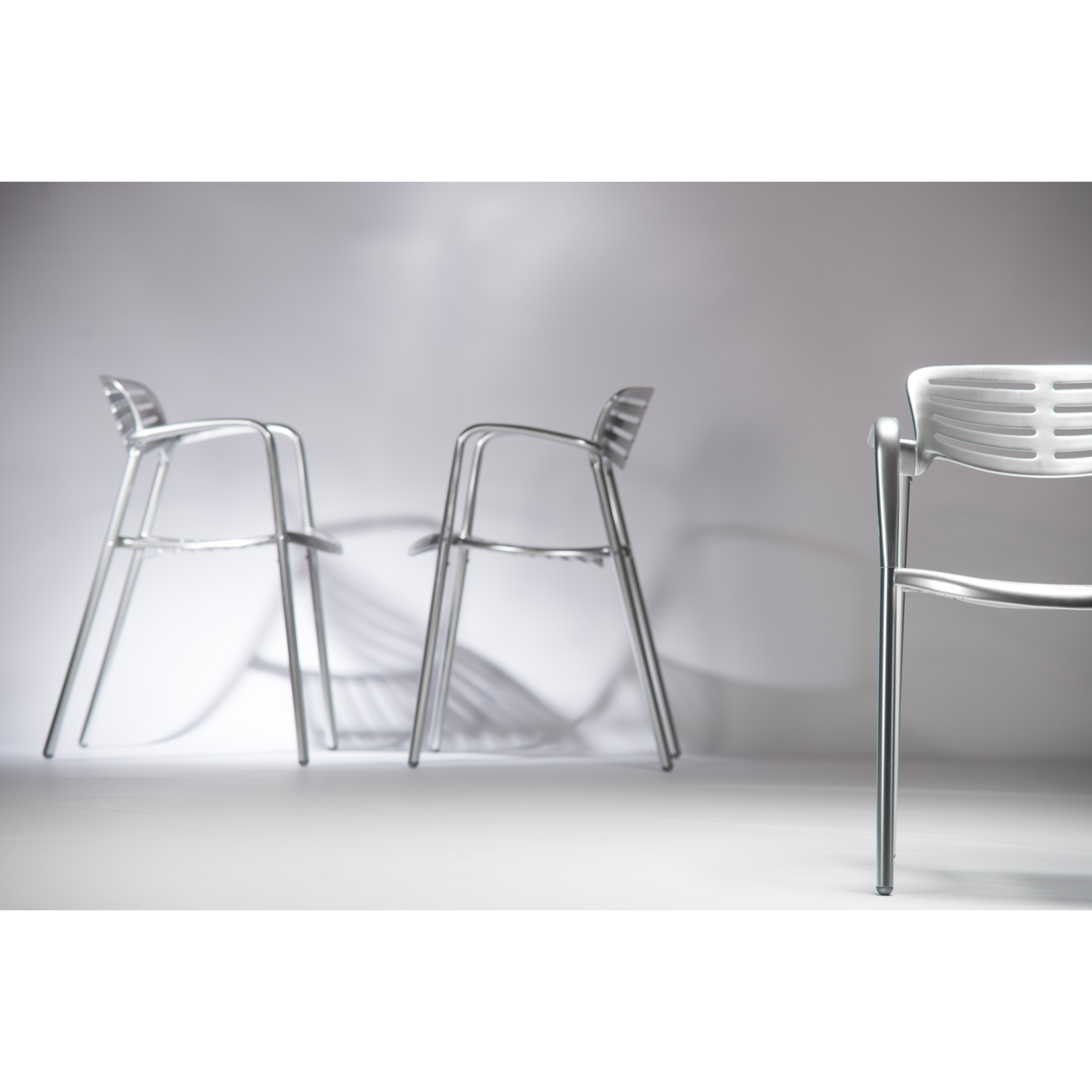 Three Toledo Armchairs by Resol, crafted with sleek curved designs, are set against a minimalist white backdrop. These UV-protected chairs, made through sustainable production methods, cast soft shadows to create an elegant and modern look.