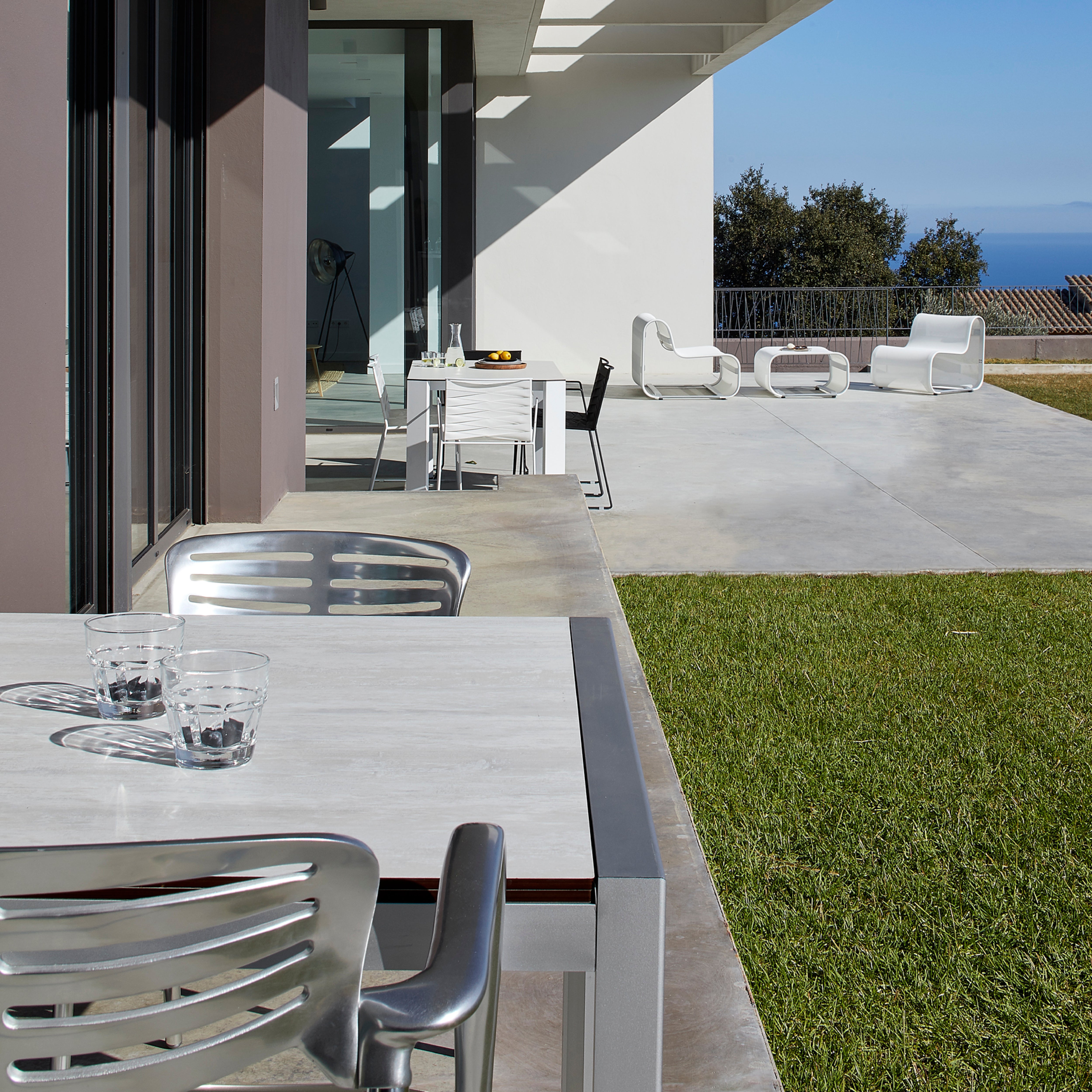 Experience the Toledo Armchair by Resol on a sustainable outdoor patio featuring a modern dining set. With glass cups and lounge chairs on concrete, enjoy ocean and tree views under UV-protected skies as light and shadows beautifully interplay.