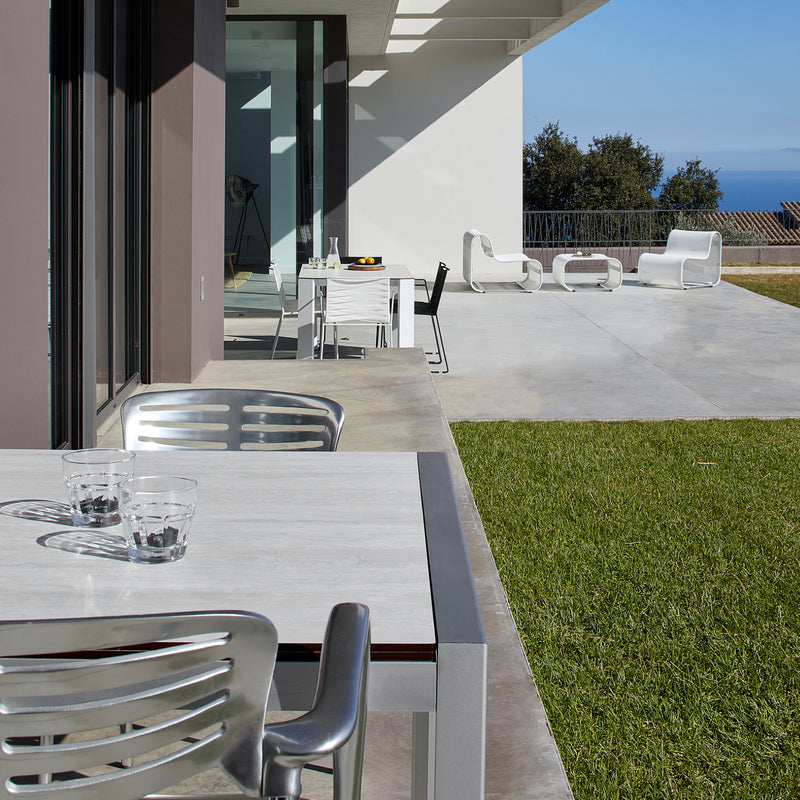Experience the Toledo Armchair by Resol on a sustainable outdoor patio featuring a modern dining set. With glass cups and lounge chairs on concrete, enjoy ocean and tree views under UV-protected skies as light and shadows beautifully interplay.