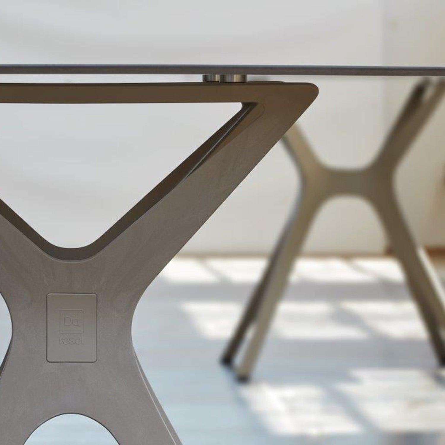 Close-up of the Vela Table S by Resol, featuring a modern wooden base with two legs forming an X and a horizontal support. A similar table leg is blurred in the background. Sunlight casts shadows on the light floor, highlighting innovative indoor and outdoor furniture solutions.