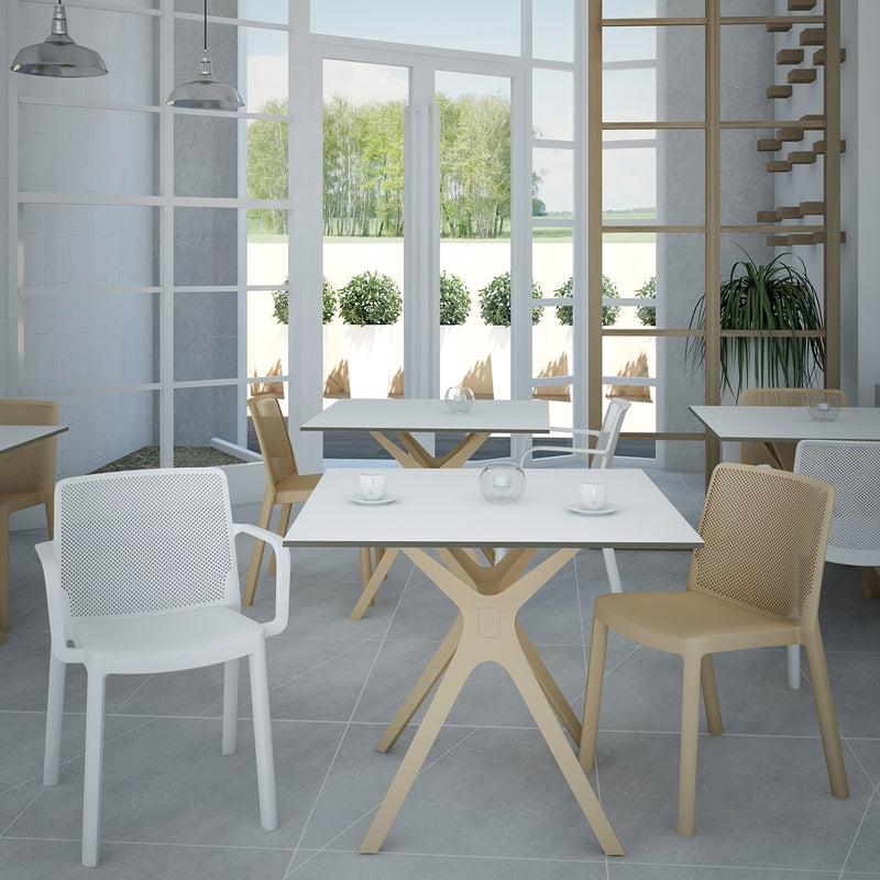 Modern café interior with minimalist design features Resols Vela Table S, surrounded by white and beige chairs. Square tables are adorned with small candles, while large windows showcase greenery and a white fence. Gray tiled floors add a sleek touch, seamlessly blurring indoor and outdoor spaces.