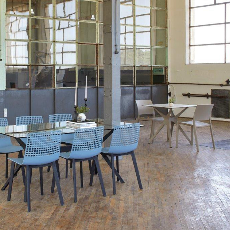 A spacious room with wooden flooring features a long glass table with light blue chairs and a smaller Vela Table S by Resol with beige chairs. Large industrial windows flood the minimalist space with natural light, highlighting versatile furniture solutions for indoor and outdoor use.