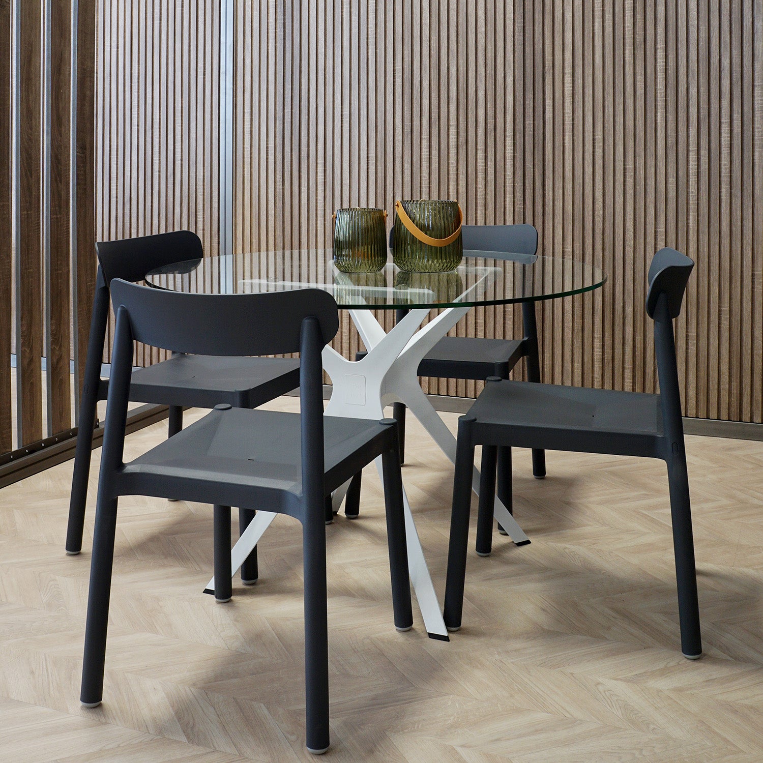 A modern dining area features a Resol Vela Table S with a round glass top and white base, complemented by four sleek black chairs. Two green glass vases are centered on the table, set against vertical wooden paneling and a light herringbone floor pattern for an innovative design.