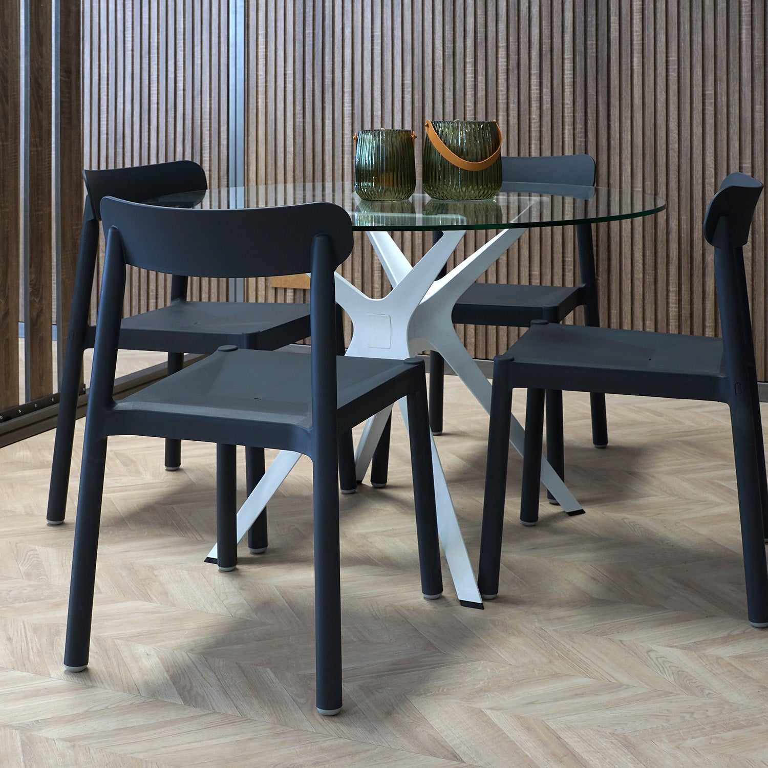 The stylish dining area showcases a Resol Vela Table S, a round glass table with a white geometric base, paired with four dark gray chairs. Set on light herringbone wood flooring against a wooden slat wall backdrop, it includes two green glass vases for both indoor and outdoor appeal.