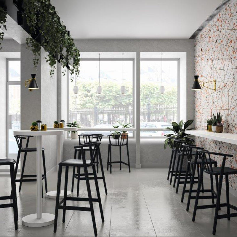 The modern cafe highlights UV-protected windows, showcasing Resols Woody Stool Upholstered at tall tables. Indoor plants enhance the sustainable vibe, while artistic patterns adorn the walls. Seamlessly blending furniture under hanging lamps creates a cozy ambiance filled with natural light.