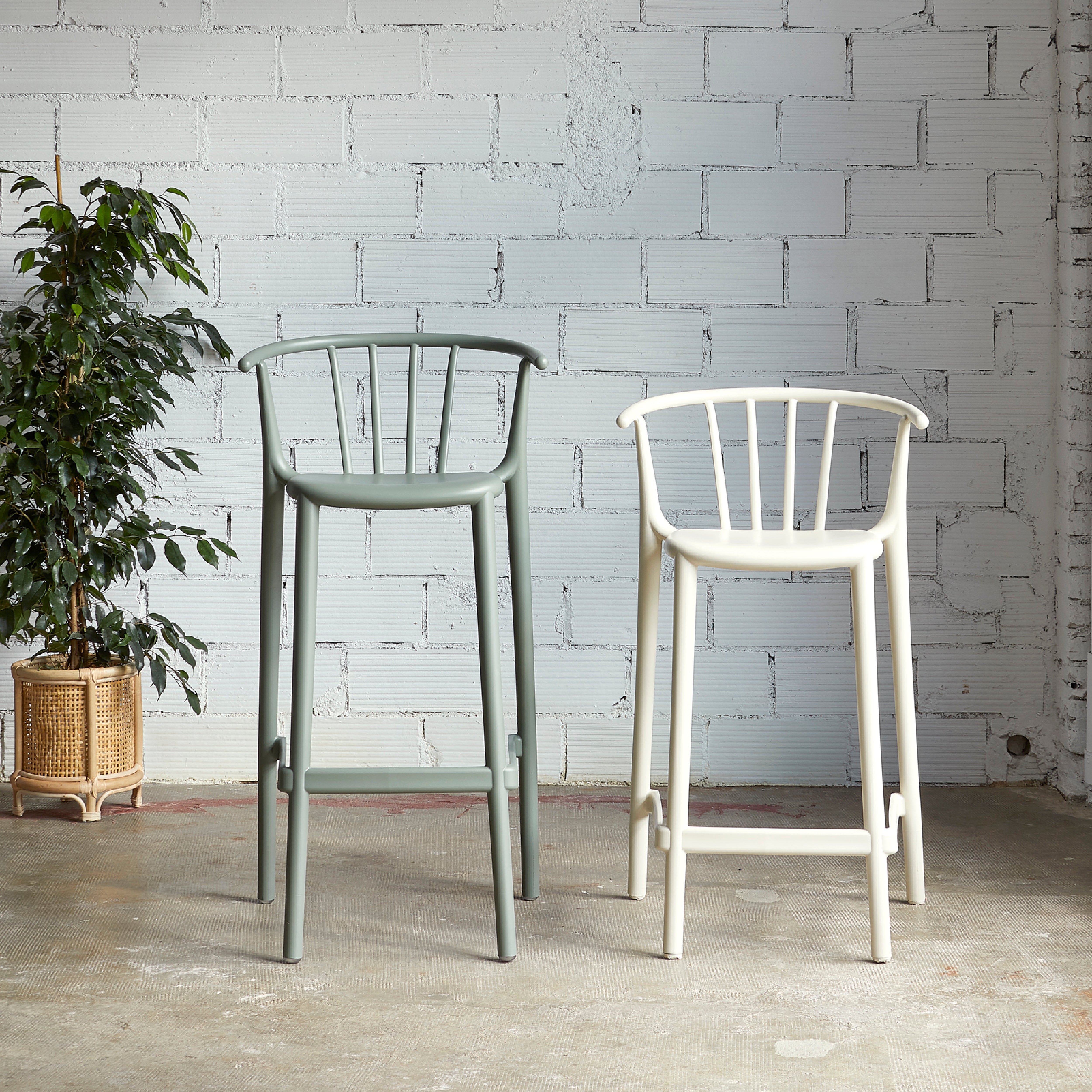 Two Resol Woody Stools, one green and one white, are placed against a white brick wall. To the left sits a potted plant in a woven basket. The concrete floor highlights the minimalist, industrial aesthetic suitable for both indoor and outdoor spaces.