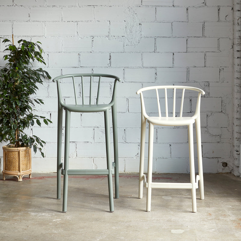 Two Resol Woody Stool Upholstered bar stools, one green and one white, stand on a concrete floor in front of a white brick wall. A leafy plant in a woven basket on the left adds sustainable production touches to the minimalist interior and exterior furniture scene.