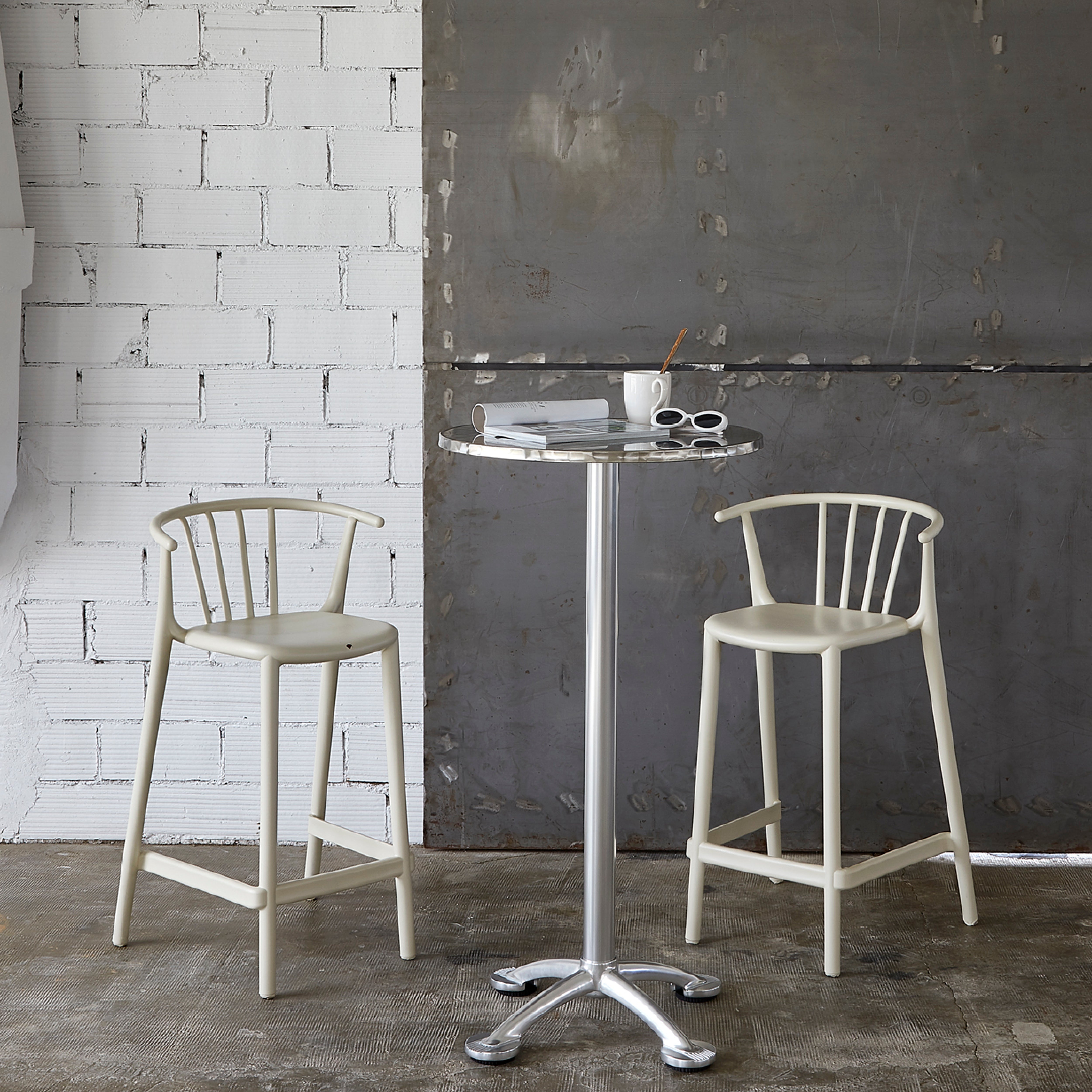 A modern, minimalistic scene showcases a tall metal table paired with two Woody Stools by Resol. The table displays a small book, white sunglasses, and a cup with a straw. A stylish textured white brick and steel panel backdrop highlights sustainable production ideals.