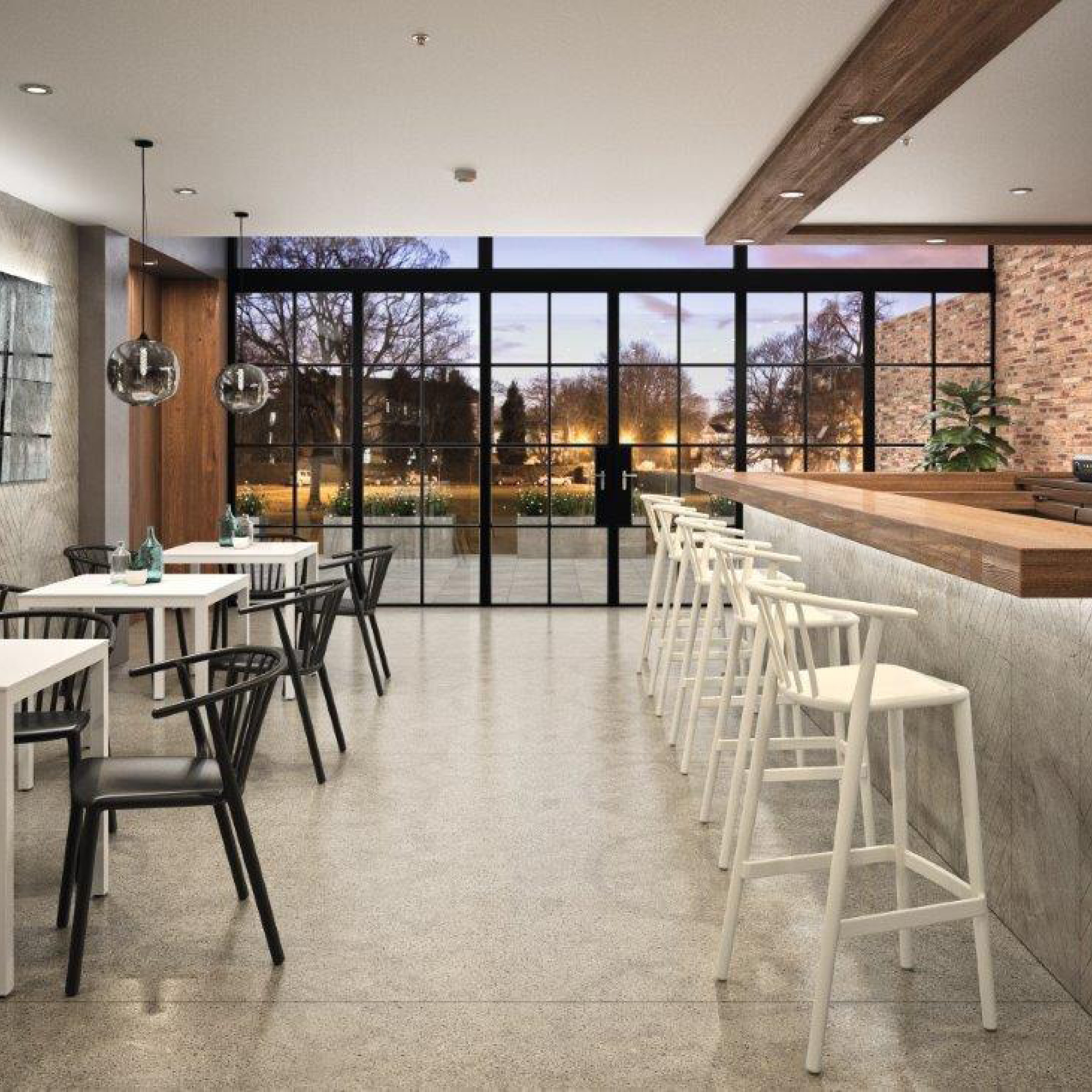 The modern café showcases Resol furniture, with a concrete floor, white tables, and black chairs to the left. On the right, a sleek bar features tall Woody Stools in white. Sustainable design is evident as large windows reveal surrounding trees and city lights while pendant lights hang overhead.