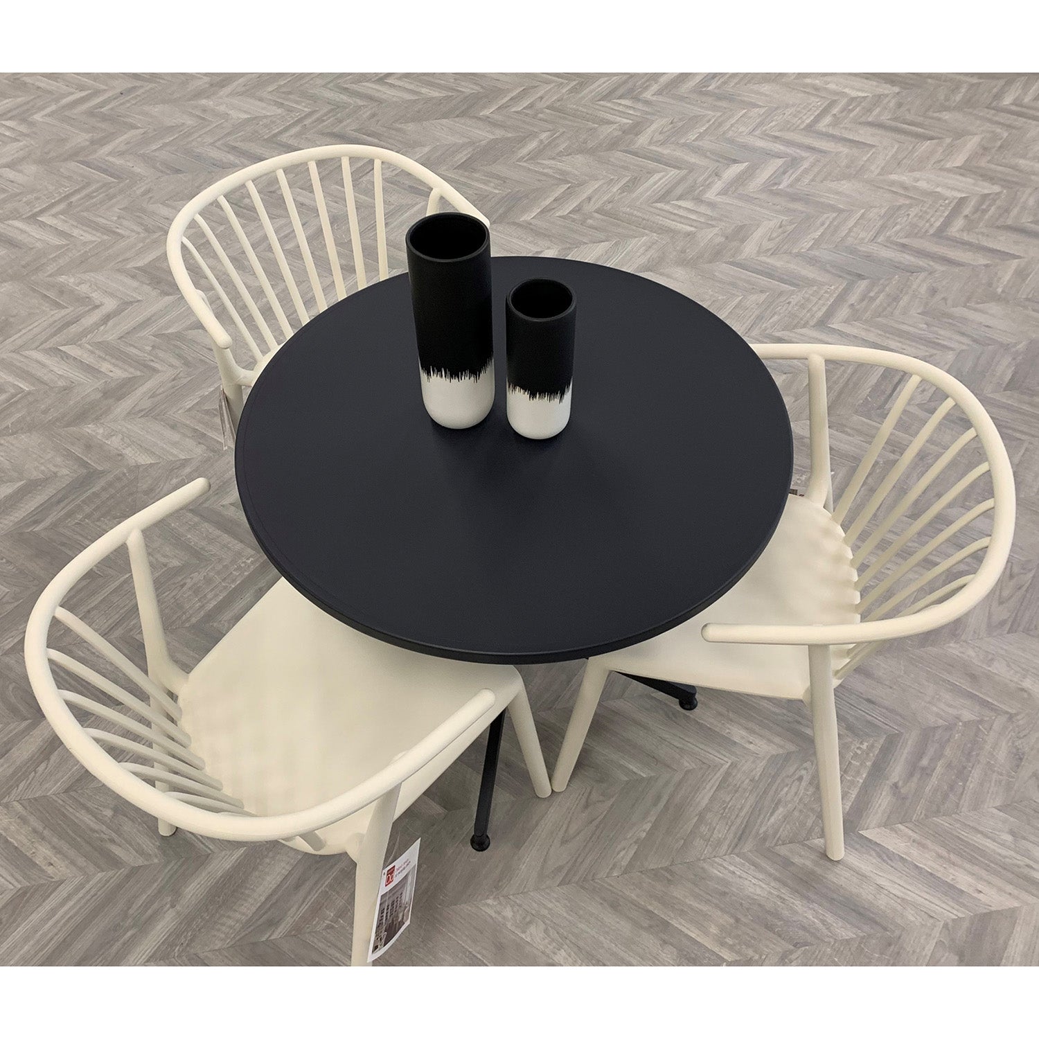 A round black UV-protected table with two black and white vases is encircled by three Resol Woody Chairs featuring curved backs, set on a light gray herringbone-patterned floor, combining elegance with sustainable production.