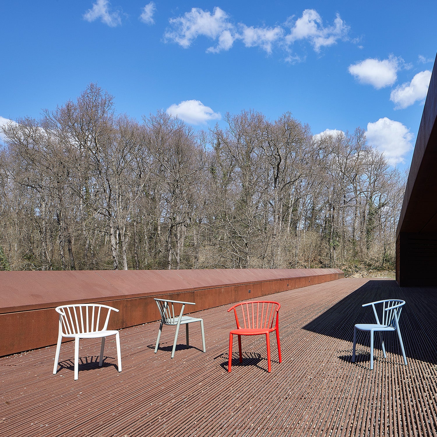Four colorful Woody Chairs by Resol—white, light green, red, and light blue—adorn a spacious outdoor wooden deck. Crafted sustainably, they complement bare trees under a clear blue sky with scattered clouds. These vibrant pieces are ideal for indoor and outdoor use.