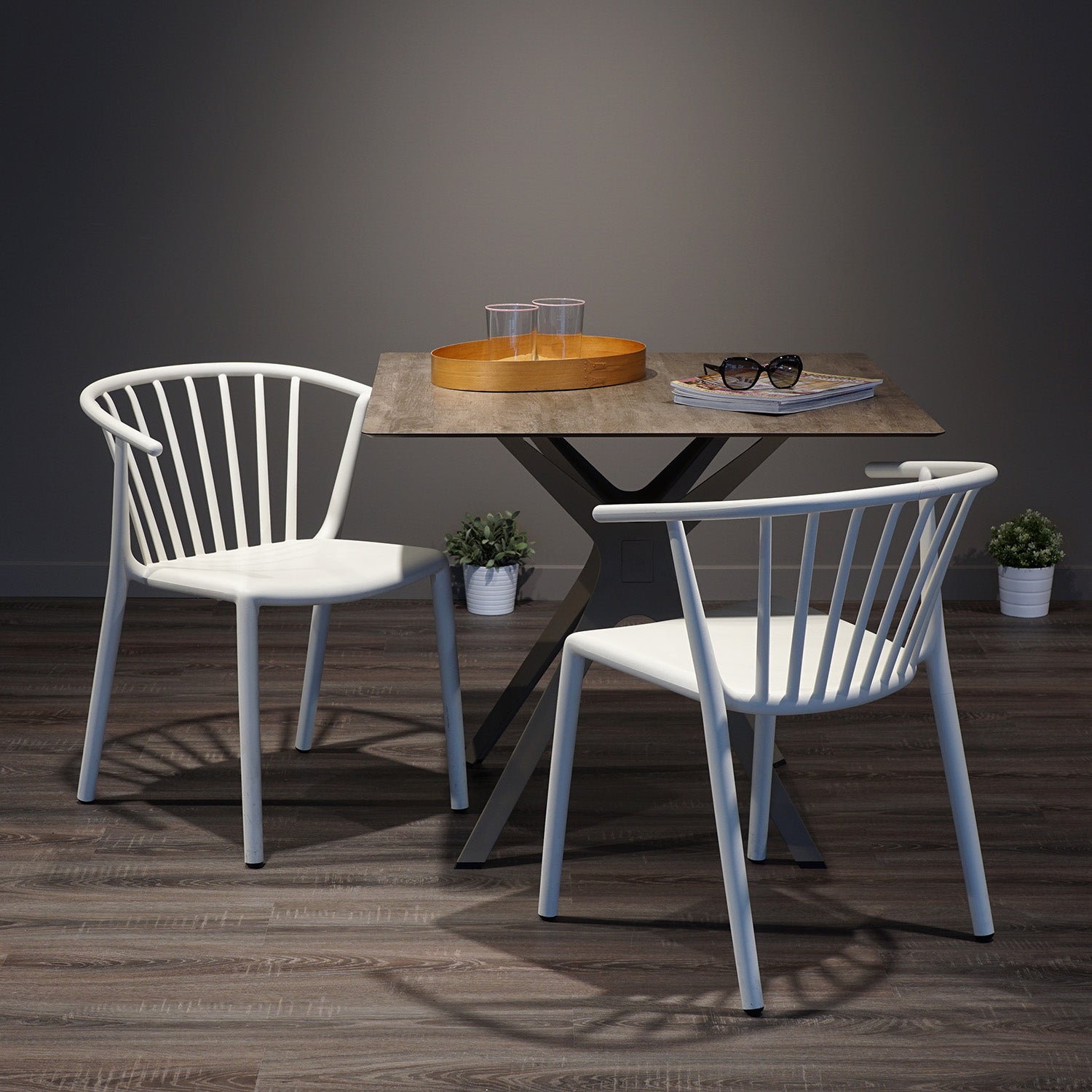 Two Resol Woody Chairs are placed around a square table with a cross-legged metal base. The tabletop features a tray, sunglasses, and magazines. UV-protected for indoor and outdoor use, this setup is enhanced by small potted plants on the wooden floor behind it.