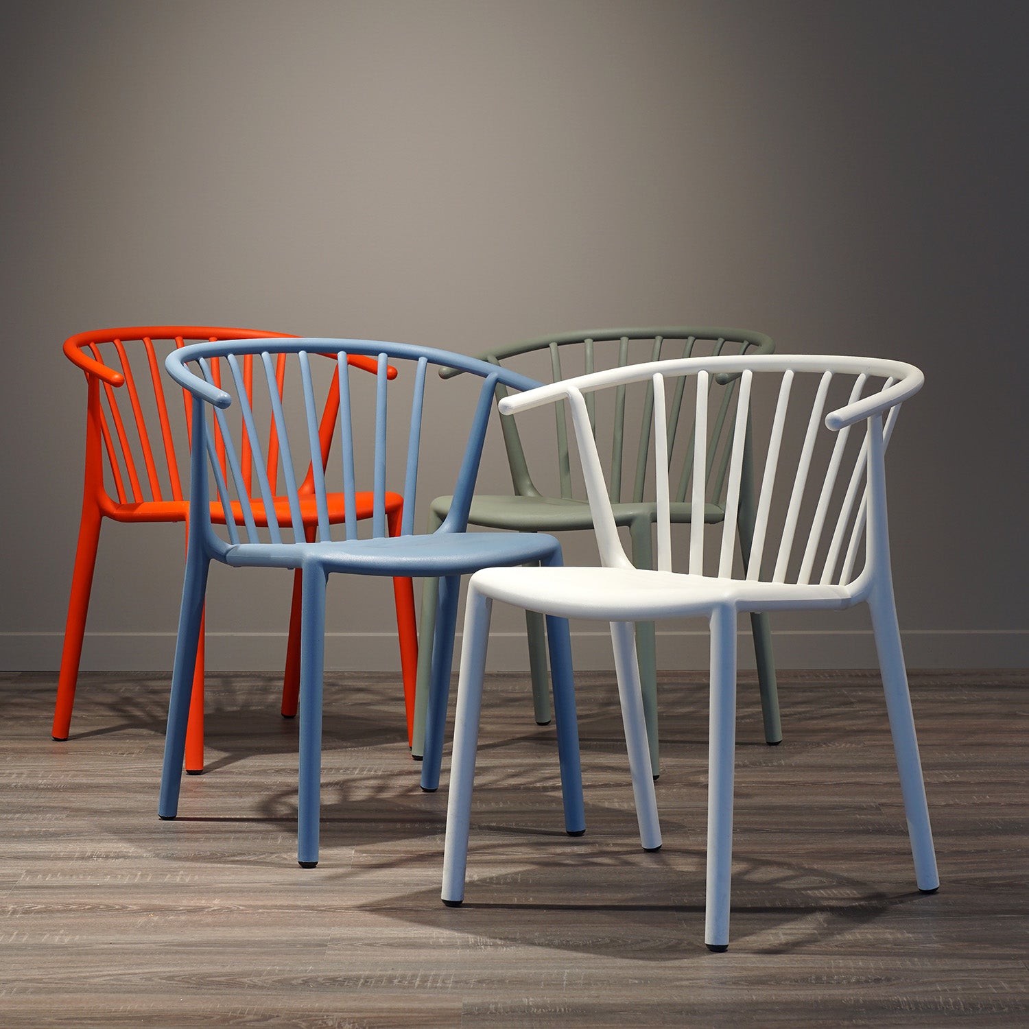 Four Woody Chairs by Resol, featuring rounded backs and open slats, are arranged on a wooden floor. Made sustainably for both indoor and outdoor use, they come in white, blue, green, and orange. Positioned under soft lighting against a plain wall.