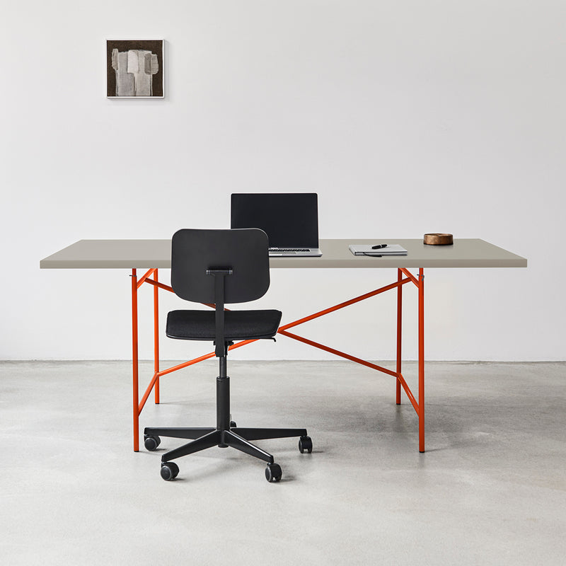 A minimalist office setup includes a Richard Lampert Eiermann 1 Table with a gray top and orange legs, paired with a black swivel chair. An open laptop, notepad, and pen sit on the table. A small abstract painting decorates the white wall above the concrete floor, enhancing the refined design.