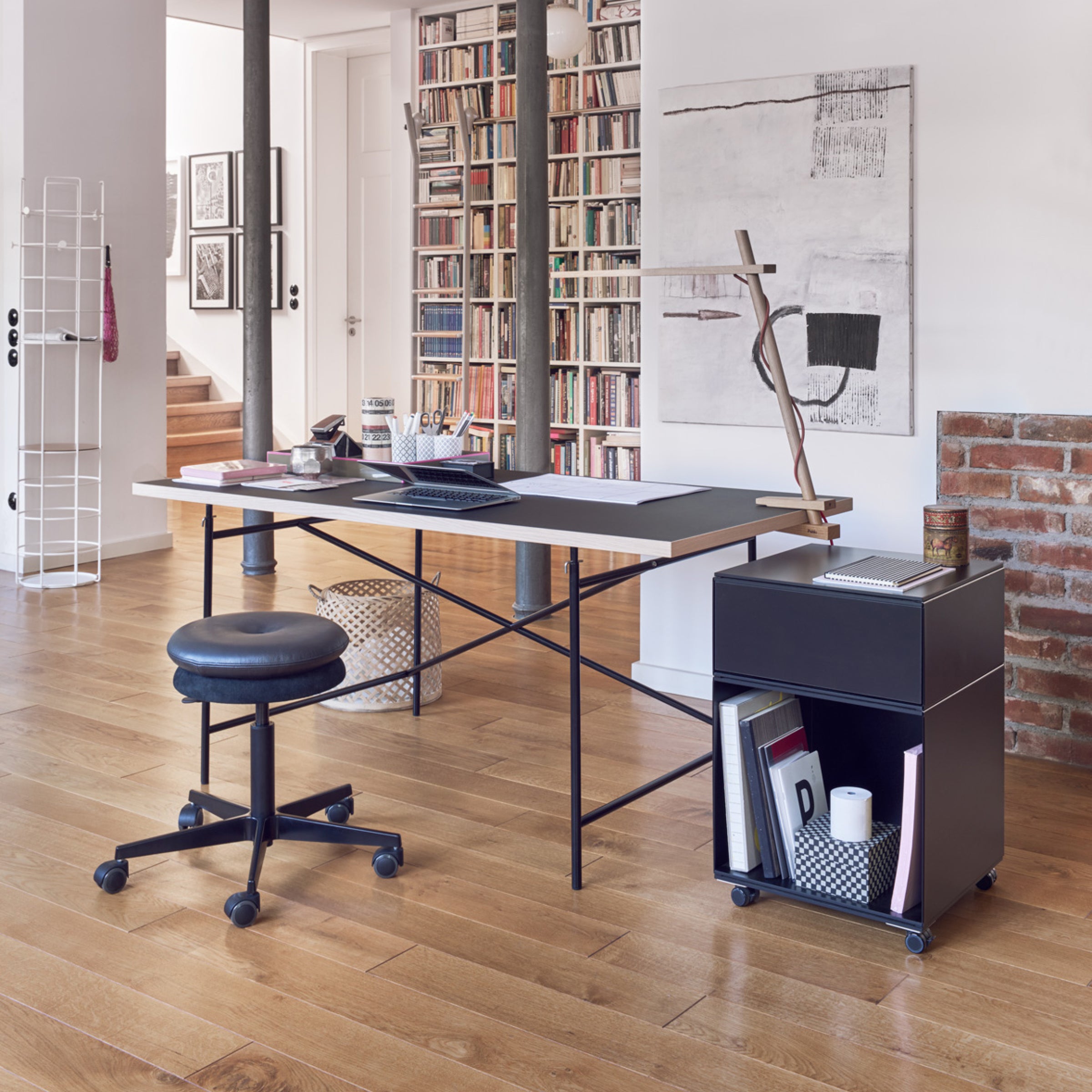 Modern home office with the Eiermann 1 Table by Richard Lampert, a black swivel chair, and a rolling storage unit. Wooden floors, bookshelves, abstract art, and exposed brick enhance the bright and organized atmosphere.