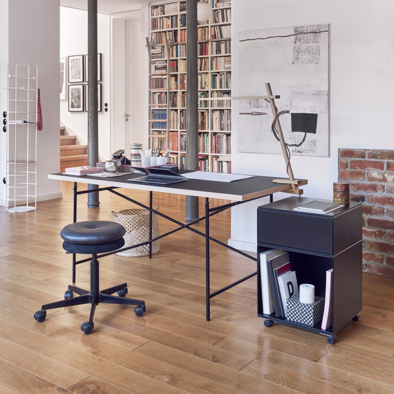 Modern home office with the Eiermann 1 Table by Richard Lampert, a black swivel chair, and a rolling storage unit. Wooden floors, bookshelves, abstract art, and exposed brick enhance the bright and organized atmosphere.