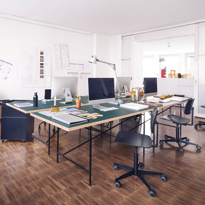 A modern office showcases a T-shaped setup with two Eiermann 1 Tables by Richard Lampert, each holding computer monitors, chairs, and office supplies. White walls, blueprints on one side, and a wooden floor enhance the clean, organized ambiance reflecting smart furniture design.