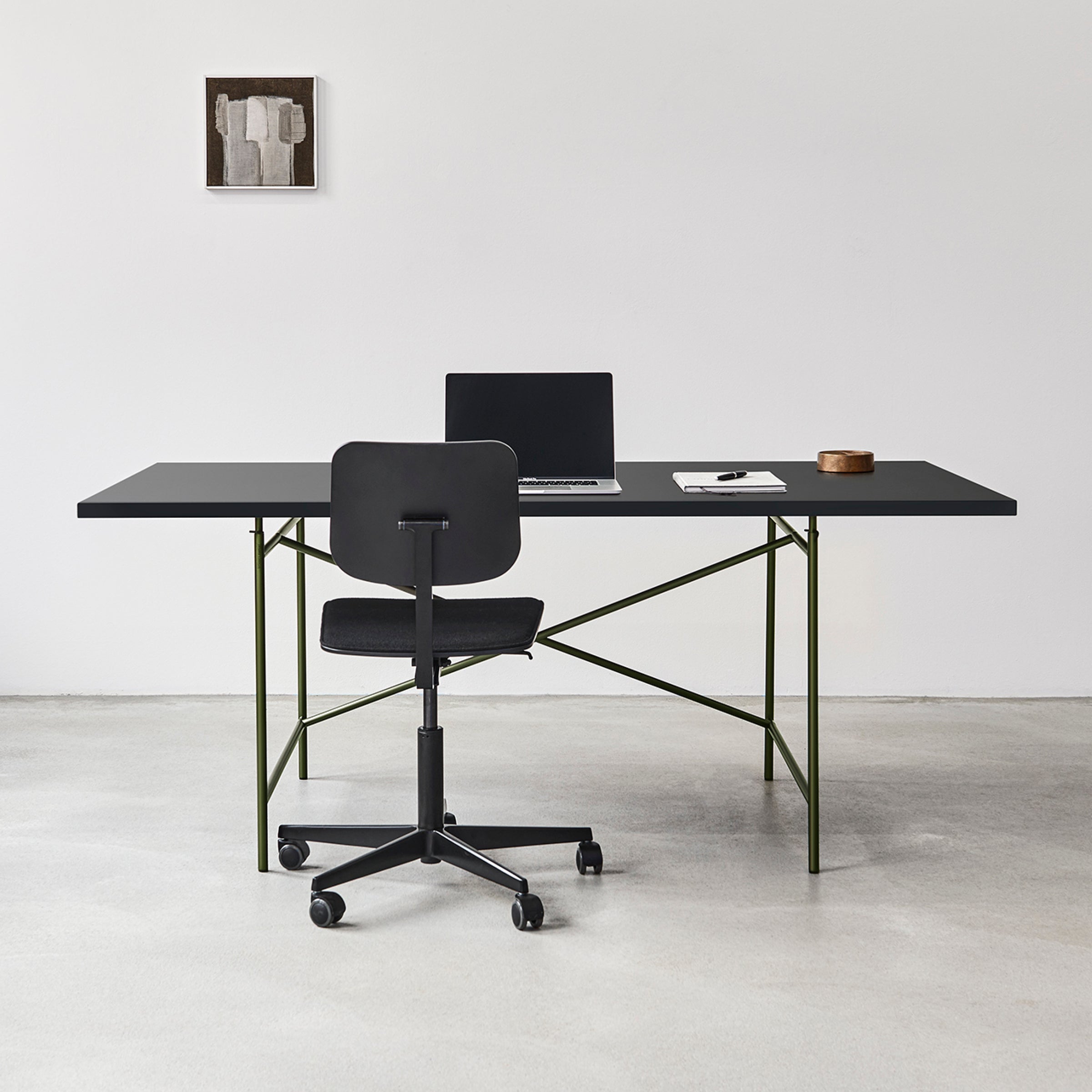 A minimalist office features the sleek Eiermann 1 Table and a matching chair on a light gray floor. An open laptop, notebook, and wooden container grace the desk. Richard Lamperts design influence is evident in the abstract art adorning the white wall.