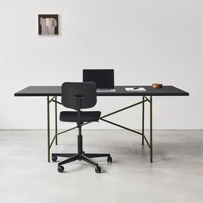 A minimalist office features the sleek Eiermann 1 Table and a matching chair on a light gray floor. An open laptop, notebook, and wooden container grace the desk. Richard Lamperts design influence is evident in the abstract art adorning the white wall.
