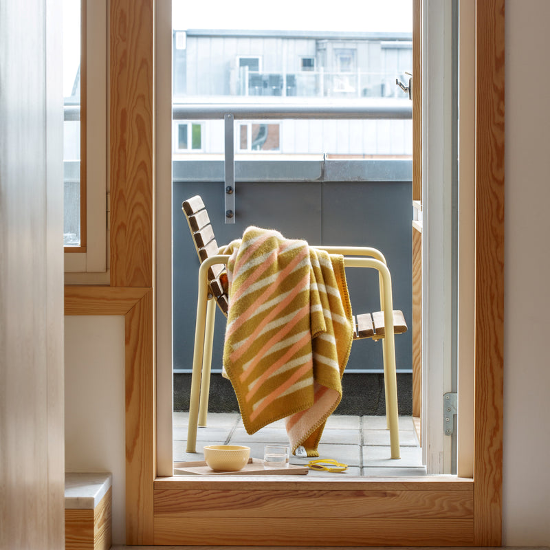 A cozy balcony scene shows a wooden chair with the Roros BISLETT Throw, made of soft Norwegian lambswool in yellow and orange stripes. A bowl and scissors are on the floor, while modern buildings dominate the view under a clear sky, evoking the mesmerizing moiré effect.